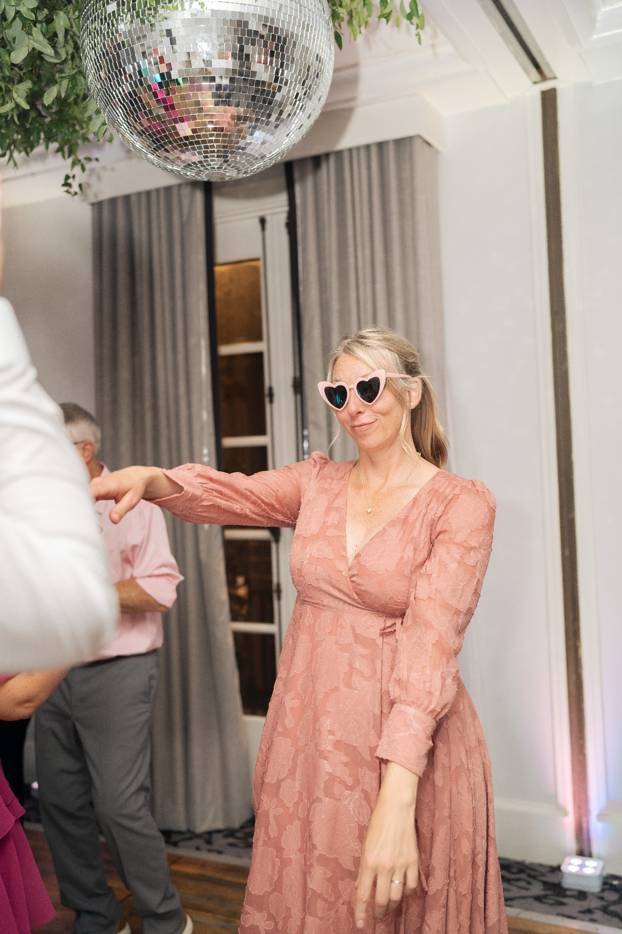 Woman in pink dress and heart-shaped sunglasses dancing under a disco ball at wedding reception
